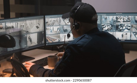 Security guard in headset controls CCTV cameras in office at night, uses computers. High tech software with surveillance cameras playback on screens. Modern security system and social safety concept.