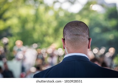 Security Guard Listening To His Earpiece On Event. Back Of Jacket Showing. Secret Service Guard. Private Bodyguard. Man With Earpiece In Crowd. Black Suit And Bald Head