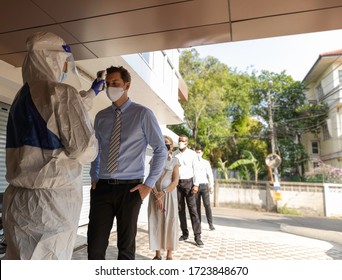 Security Guard In PPE Suit Uses Infrared Thermometer Measuring Temperature With European Staff Scanning For Coronavirus Or Covid-19 Symptom At Office Entrance. International Medical Healthcare System