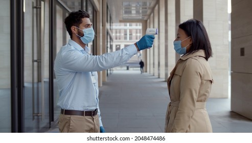 Security Guard In Safety Mask Standing Outside Business Center And Check Body Temperature Of Staff. Businesswoman Scan Temperature With Infrared Thermometer Outside Office Building