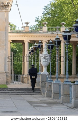 Security guard in uniform walks on pathway during safety patrol monitoring in historic palace park. Male watchman on duty represents law enforcement officer, public surveillance, and protection.