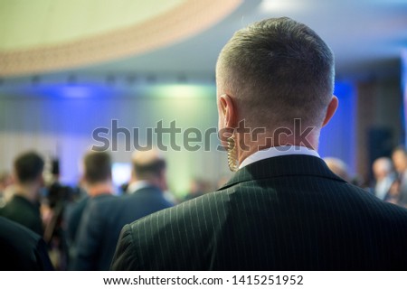 Security on public event. Secret guard service. Private bodyguard with earpiece standing among crowd. Safety of govern and business meeting. Secret service agent listening to his earpiece, side.
