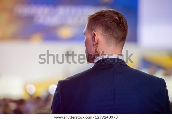 Security on public event. Secret guard service. \
Private bodyguard with earpiece standing among crowd. Safety of\
govern and business meeting. Secret service agent listening to his\
earpiece, side.