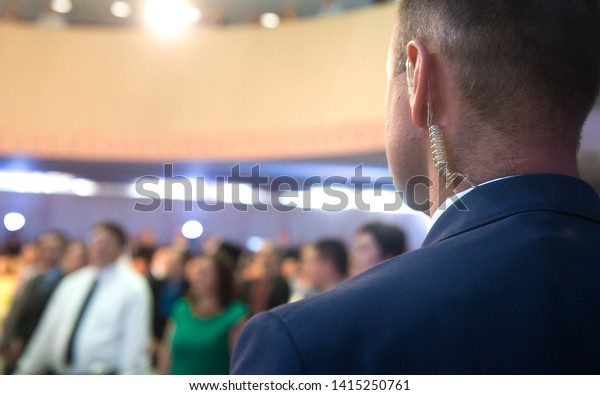 Security on public event. Secret guard service.\
Private bodyguard with earpiece standing among crowd. Safety of\
govern and business meeting. Secret service agent listening to his\
earpiece, side.