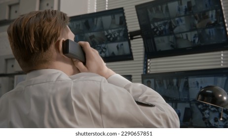 Security operator talks by landline phone in observation room. Security officer monitors CCTV cameras on computer. Screens showing security cameras footage with facial recognition software. Back view. - Shutterstock ID 2290657851