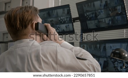 Security operator talks by landline phone in observation room. Security officer monitors CCTV cameras on computer. Screens showing security cameras footage with facial recognition software. Back view.
