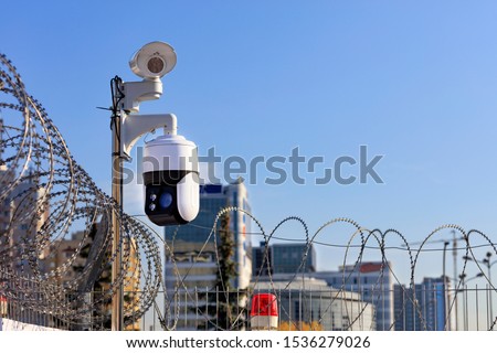 Security perimeter. Barbed wire fencing, a red light and cameras protect against intrusions against the background of urban buildings and the blue sky in blur.