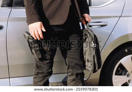 A security professional in tactical gear holding a briefcase and standing next to a car. The holstered firearm and formal attire convey themes of personal protection, security, and professional duty.