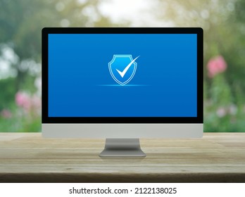 Security shield with check mark flat icon on desktop modern computer monitor screen on wooden table over blur pink flower and tree in park, Technology internet cyber security and anti virus online