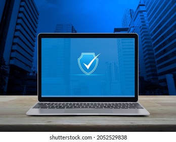 Security Shield With Check Mark Icon On Modern Laptop Computer Monitor Screen On Wooden Table Over Office City Tower And Skyscraper, Technology Internet Cyber Security And Anti Virus Online Concept