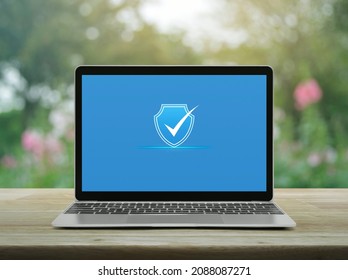 Security shield with check mark icon on modern laptop computer monitor screen on wooden table over blur pink flower and tree in park, Technology internet cyber security and anti virus online concept