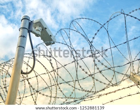 security or surveillance camera hanging behind barbed wire fence as a wall in a controlled place or prison with a blue sky background, security concept  