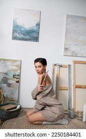 Seductive brunette artist holding paintbrushes while sitting on floor in workshop - Shutterstock ID 2250184401