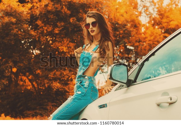 seductive lady with big silicon breasts\
wears jeans overall and sits on a car in the\
autumn