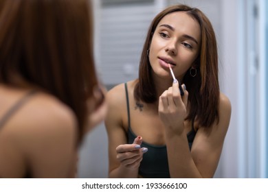 Seductive pretty young woman using lip brush for applying red lipstick to her lips looking in mirror - Powered by Shutterstock