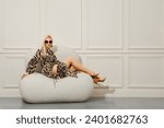 Seductive woman with bare long legs in sandals sits in her living room, full-length on a plush armchair