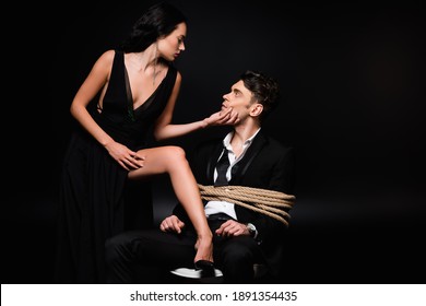 Seductive Woman In Dress Touching Face Of Tied Submissive Man Sitting On Chair On Black