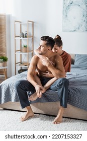 Seductive Woman Hugging Shirtless Man In Pajama Pants Sitting On Bed And Looking Away