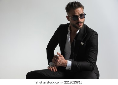 Seductive Young Man In Black Tuxedo Holding Elbow On Knee, Looking Away And Posing In Front Of Grey Background In Studio