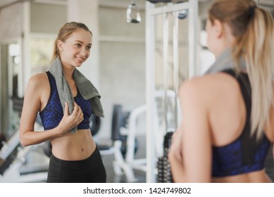 See From Back Side Of Young Sport Caucasian Slim Woman Is Looking Herself Big Mirror At Gym. Woman Workout And Training At Fitness Room Concept.