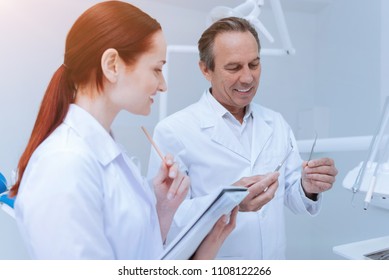 See The Difference. Smiling Male Person Wearing Clean Smock Demonstrating Two Medical Tools While Looking Downwards