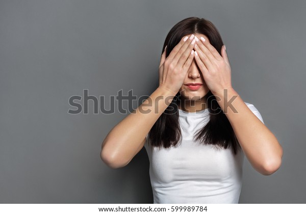 See no evil
concept. Portrait of young scared woman covering eyes with hands
while standing against gray studio background. Confused girl close
eyes with palms ignoring
something