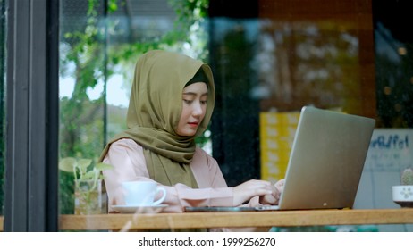 See Through Clear Glass,  Portrait Young Pretty Muslim Woman Typing On Laptop And Looking Screen While Working In Coffee Shop With Happiness, Muslim Business Working Concept