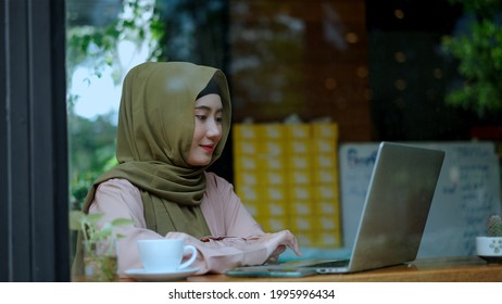 See Through Clear Glass,  Young Pretty Muslim Woman Typing On Laptop And Looking Screen And Smile While Working In Coffee Shop With Happiness, Muslim Business Working Concept