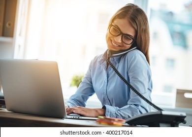 See You Tomorrow! Cheerful Young Beautiful Woman In Glasses Talking On The Phone And Using Laptop With Smile While Sitting At Her Working Place