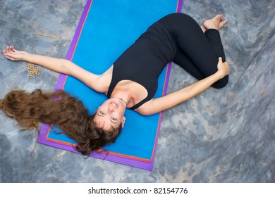 Seen from above, a brown haired caucasian woman doing yoga exercise, Ardha Jathara Parivarttanasana Pose or Half Revolved Belly posture  on yoga mat in studio with mottled background.