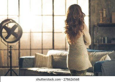 Seen From Behind, A Brunette Woman In Comfortable Clothing Is Standing In A Loft Living Room, Looking Out The Loft Window. Urban Chic Loft Decoration Details.