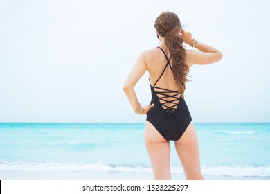 Seen From Behind Elegant 40 Year Old Woman With Long Curly Hair In Elegant Black Swimsuit On A White Beach Looking Into The Distance.