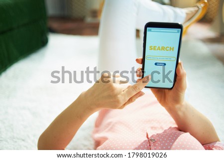 Seen from behind female in blouse and white pants in the living room in sunny day searching for carpet cleaning service on a smartphone while laying on white carpet.