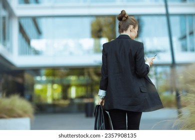 Seen from behind middle aged business woman near office building in black jacket with smartphone and briefcase walking.
