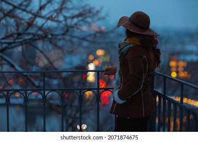 Seen From Behind Middle Aged Woman In Brown Hat And Scarf Outside In The City Park In Winter In Sheepskin Coat At Night.