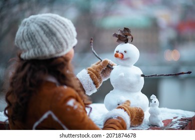Seen from behind middle aged woman with mittens in a knitted hat and sheepskin coat making a snowman outside in the city park in winter.