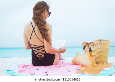 Seen From Behind Modern 40 Year Old Woman With Long Curly Hair In Elegant Black Swimsuit On A White Beach Reading Book.