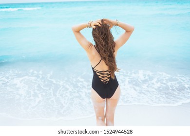 Seen From Behind Modern 40 Year Old Woman With Long Curly Hair In Elegant Black Swimsuit On A White Beach Walking Towards Sea.