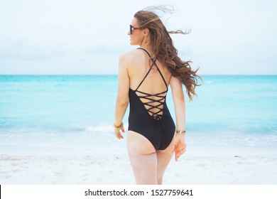 Seen From Behind Modern 40 Year Old Woman With Long Curly Hair In Elegant Black Bathing Suit On A White Beach Walking Towards Sea.