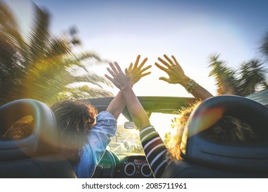 Seen From Behind A Pair Of Euphoric Women In A Convertible Car, Twisting And Waving. Two Curly Girls On Vacation, Having Fun In Topless Auto. Female Arms Up, Palms Open Driving In The Sunny Street