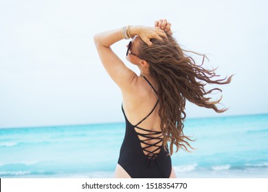 Seen From Behind Stylish 40 Year Old Woman With Long Curly Hair In Elegant Black Swimsuit On A White Beach Relaxing.