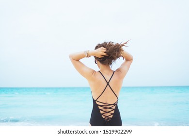 Seen From Behind Stylish 40 Year Old Woman With Long Curly Hair In Elegant Black Swimsuit On A White Beach Adjusting Hair.