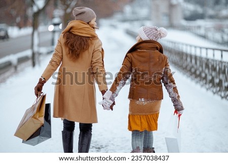 Seen from behind stylish mother and child in coat, hat, scarf and mittens with shopping bags walking outdoors in the city in winter.