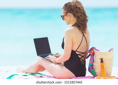 Seen From Behind Trendy 40 Year Old Woman With Long Curly Hair In Elegant Black Bathing Suit On A White Beach Working On A Laptop.