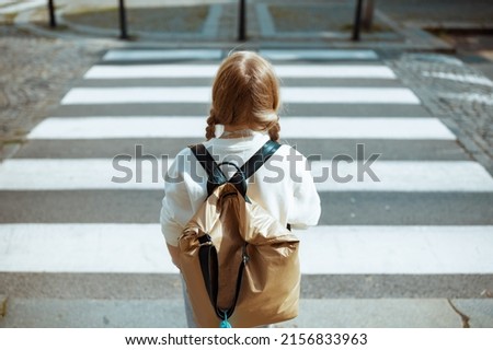 Seen from behind trendy pupil in white sweatshirt with backpack crossing crosswalk and going to school outdoors in the city.