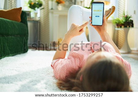 Seen from behind woman in blouse and white pants in the living room in sunny day searching for carpet cleaning service on a smartphone while laying on white carpet.