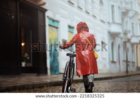 Seen from behind woman in red rain coat with bicycle walking in the rain outside in the city.