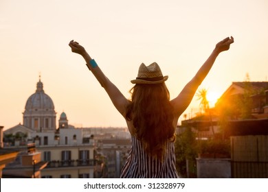 Seen From Behind, A Woman Is Standing With Outstretched Arms, Looking Out At The City Of Rome At Sunset In Summer.