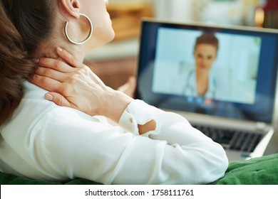 Seen From Behind Woman In White Blouse With Neck Pain Speaking With Doctor Using Tele Health Technology In The Living Room In Sunny Day.