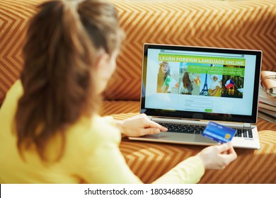 Seen from behind woman in yellow jacket with credit card subscribing on learning foreign language online site on a laptop at home in sunny day.
